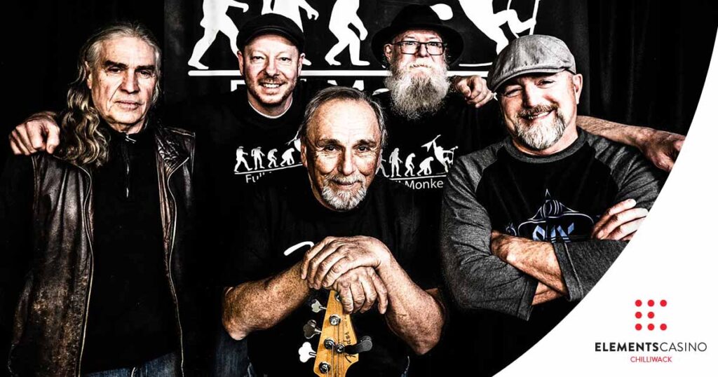 Five men pose together, smiling, standing in front of a dark backdrop at Elements Casino Chilliwack. One man holds a guitar, and the Elements Casino Chilliwack logo appears in the bottom right corner.