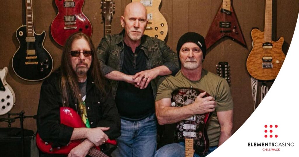 Three men holding electric guitars pose together in front of a wall displaying several hanging guitars at Elements Casino Chilliwack. The man in the middle stands while the others sit, with the Elements Casino Chilliwack logo appearing in the lower right corner.
