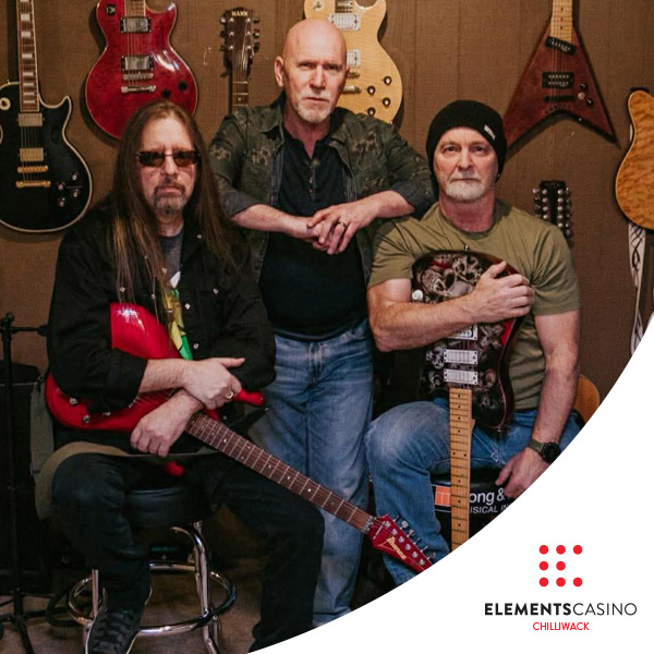 Three men with guitars pose in front of a wall displaying even more guitars. The man in the center stands, while the others sit holding electric guitars. The Elements Casino Chilliwack logo is visible at the bottom right corner, highlighting the venue’s vibrant musical atmosphere.