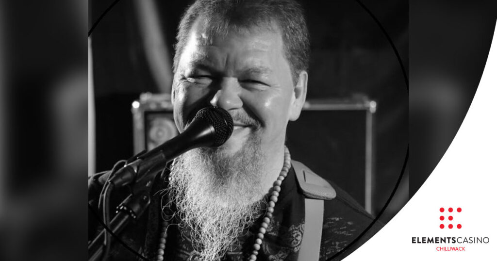 A smiling man with a long beard sings into a microphone; he wears a necklace and holds a guitar. The image is black and white, capturing the lively spirit of live music at Elements Casino Chilliwack. The Elements Casino Chilliwack logo is in the bottom right corner.