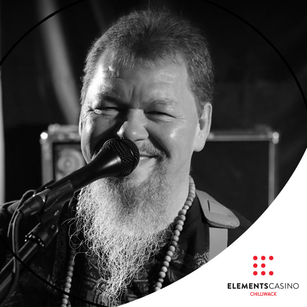 A bearded man smiling and singing into a microphone on stage, with a guitar strap over his shoulder. The image is black and white, with the Elements Casino Chilliwack logo appearing in the corner, capturing the lively atmosphere of a performance at Elements Casino Chilliwack.