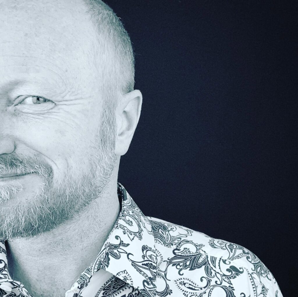 Close-up portrait of a smiling man with a beard wearing a patterned shirt against a black background.